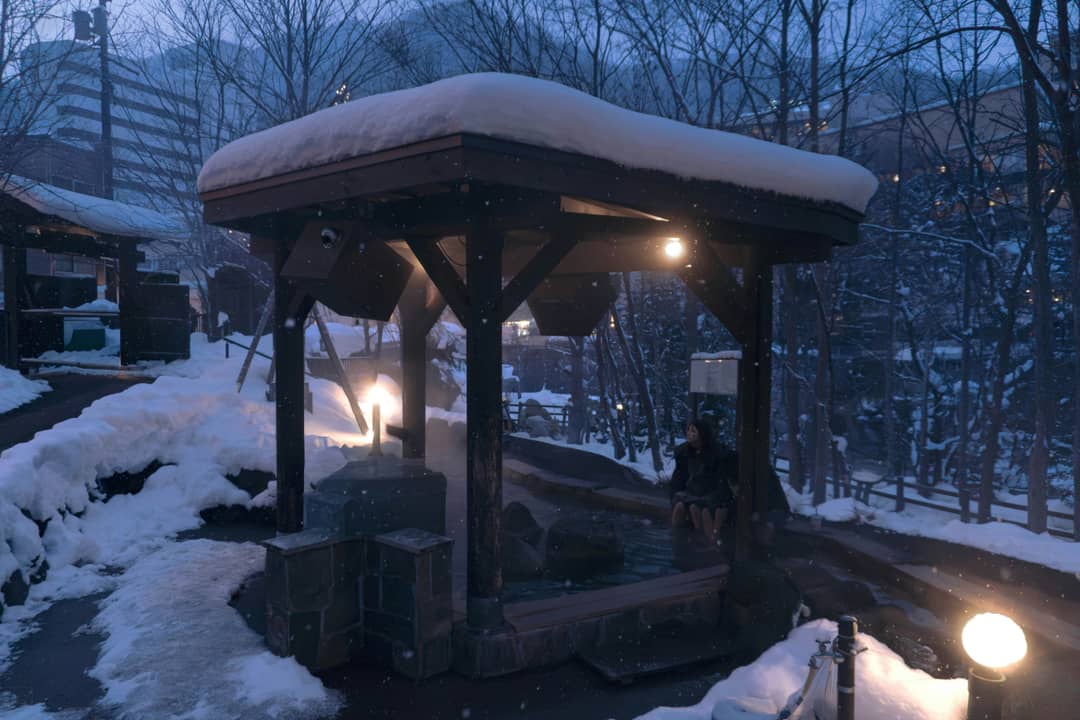 雪の降る夕暮れ、湯小屋の明かりだけが灯る温泉街の一角